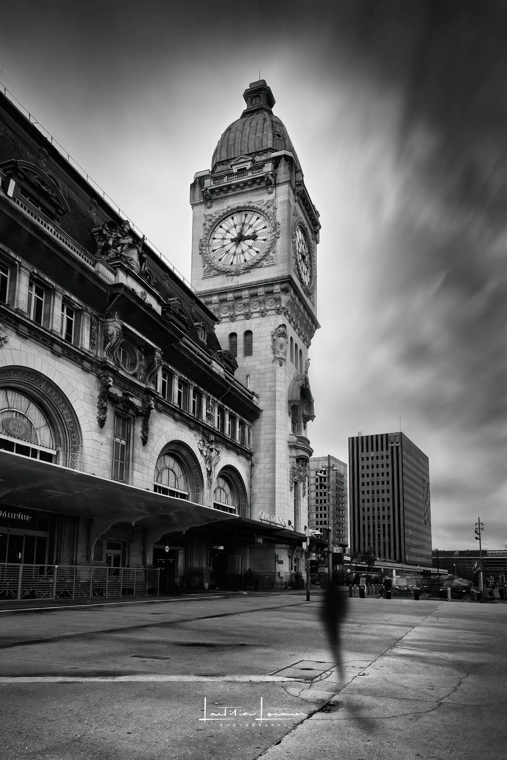 Photographie en noir et blanc de la Gare de Lyon à Paris, capturée en pose longue avec des silhouettes de passants floutées et un jeu de contrastes.