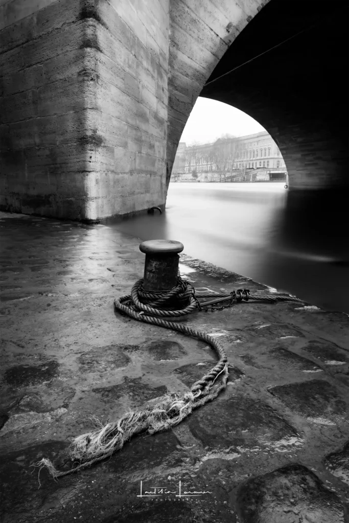 Photographie en pose longue sous un pont de Paris, avec une corde usée et le reflet de la Seine. Noir et blanc.